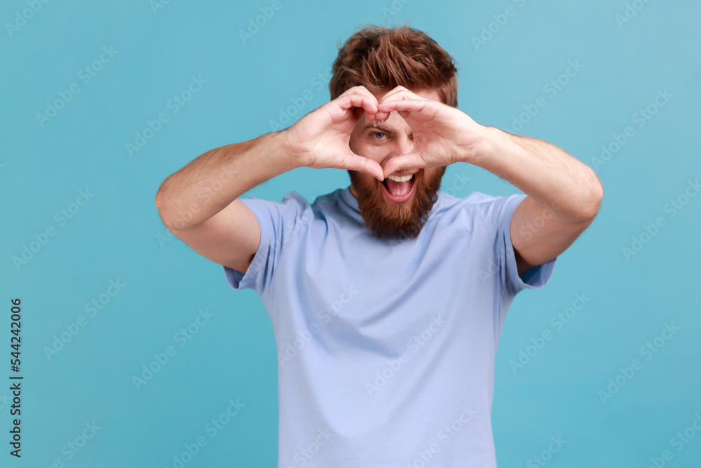 Portrait of handsome bearded man looking through hands in shape of heart showing romantic gesture, love confession, valentines day celebration. Indoor studio shot isolated on blue background. Portrait of handsome bearded man looking through hands in shape of heart showing romantic gesture, love confession, valentines day celebration. Indoor studio shot isolated on blue background.