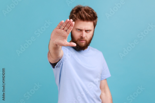 Portrait of strict bearded man showing stop sign at camera, standing with frowning face and bossy expression, meaning caution to avoid danger or mistake. Indoor studio shot isolated on blue background