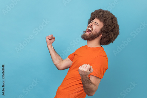 Fotografija Side view of excited positive happy man with Afro hairstyle wearing orange T-shirt screaming for joy with raised fists and closed eyes, rejoicing win