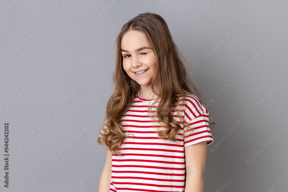 Portrait of playful cheerful little girl wearing striped T-shirt standing and winking playfully, positive expression, looking at camera. Indoor studio shot isolated on gray background. Portrait of playful cheerful little girl wearing striped T-shirt standing and winking playfully, positive expression, looking at camera. Indoor studio shot isolated on gray background.