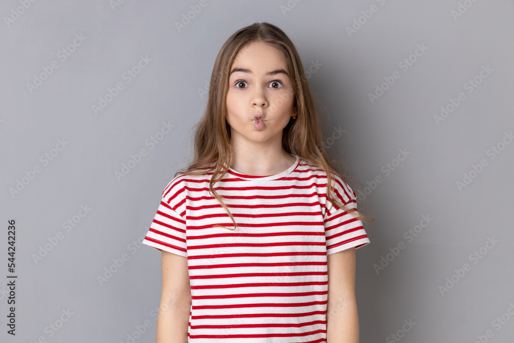 Funny surprised little girl wearing striped T-shirt making fish face with pout lips and looking amazed, choking with idiotic comical silly expression. Indoor studio shot isolated on gray background. Funny surprised little girl wearing striped T-shirt making fish face with pout lips and looking amazed, choking with idiotic comical silly expression. Indoor studio shot isolated on gray background.