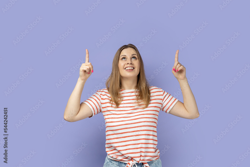 Look there, advertising area. Portrait of smiling blond woman wearing striped T-shirt looking astonished and pointing up at empty space for promotion. Indoor studio shot isolated on purple background. Look there, advertising area. Portrait of smiling blond woman wearing striped T-shirt looking astonished and pointing up at empty space for promotion. Indoor studio shot isolated on purple background.