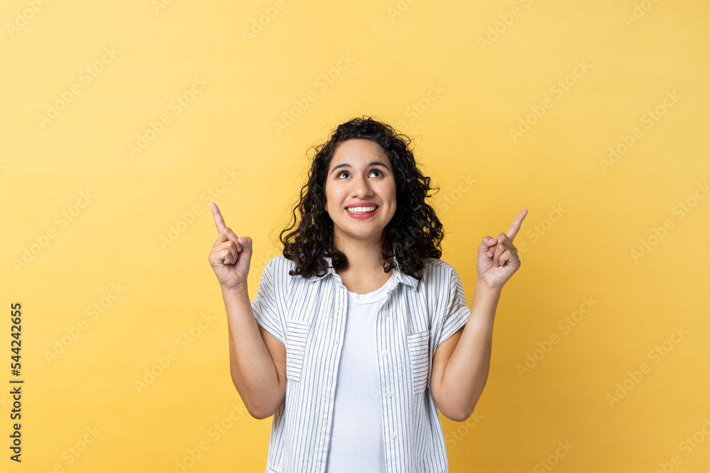 Portrait of satisfied delighted woman with dark wavy hair pointing up at empty place for ad content and expressing positive emotions. Indoor studio shot isolated on yellow background. Portrait of satisfied delighted woman with dark wavy hair pointing up at empty place for ad content and expressing positive emotions. Indoor studio shot isolated on yellow background.