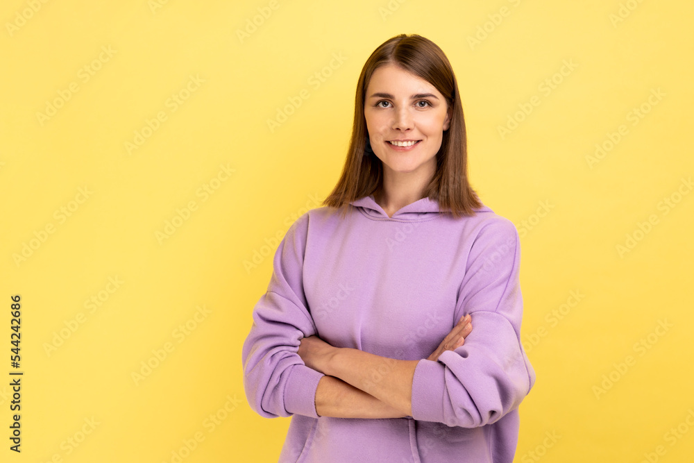 Portrait of happy successful woman standing with crossed hands, looking at camera with charming and joyous toothy smile, wearing purple hoodie. Indoor studio shot isolated on yellow background. Portrait of happy successful woman standing with crossed hands, looking at camera with charming and joyous toothy smile, wearing purple hoodie. Indoor studio shot isolated on yellow background.