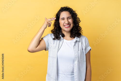 Portrait of woman with dark wavy hair pointing finger gun to head and looking desperate, making suicide gesture, shooting herself. Indoor studio shot isolated on yellow background.