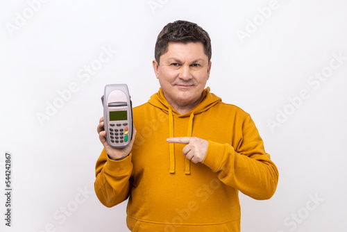 Smiling positive man holding and pointing at pos payment terminal, using paypass for purchases payment, wearing urban style hoodie. Indoor studio shot isolated on white background. photo