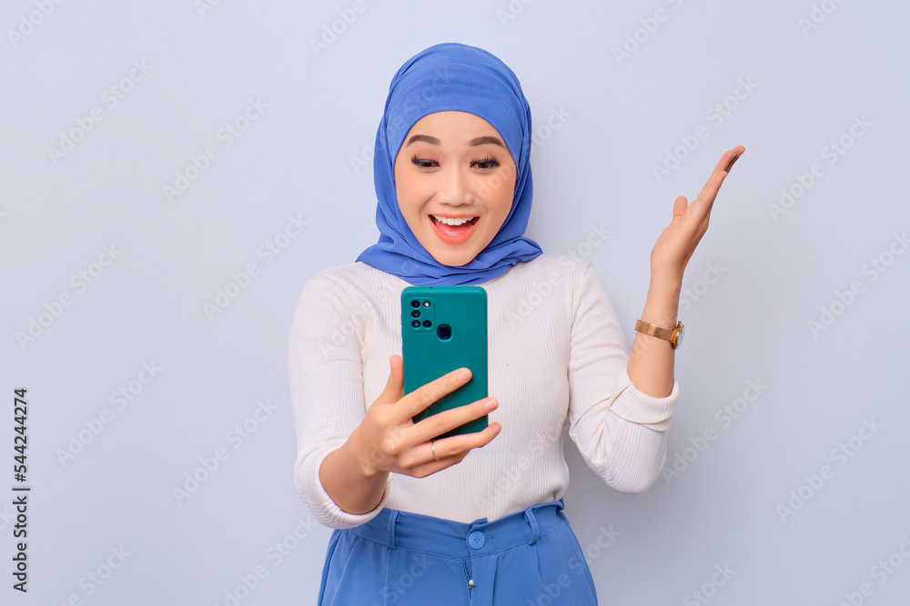 Excited young Asian Muslim woman looking at smartphone getting good news isolated over white background