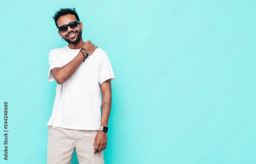 Handsome hipster smiling model. Sexy unshaven man dressed in white summer t-shirt and jeans clothes. Fashion male posing near blue wall in studio. Isolated. In sunglasses Handsome hipster smiling model. Sexy unshaven man dressed in white summer t-shirt and jeans clothes. Fashion male posing near blue wall in studio. Isolated. In sunglasses