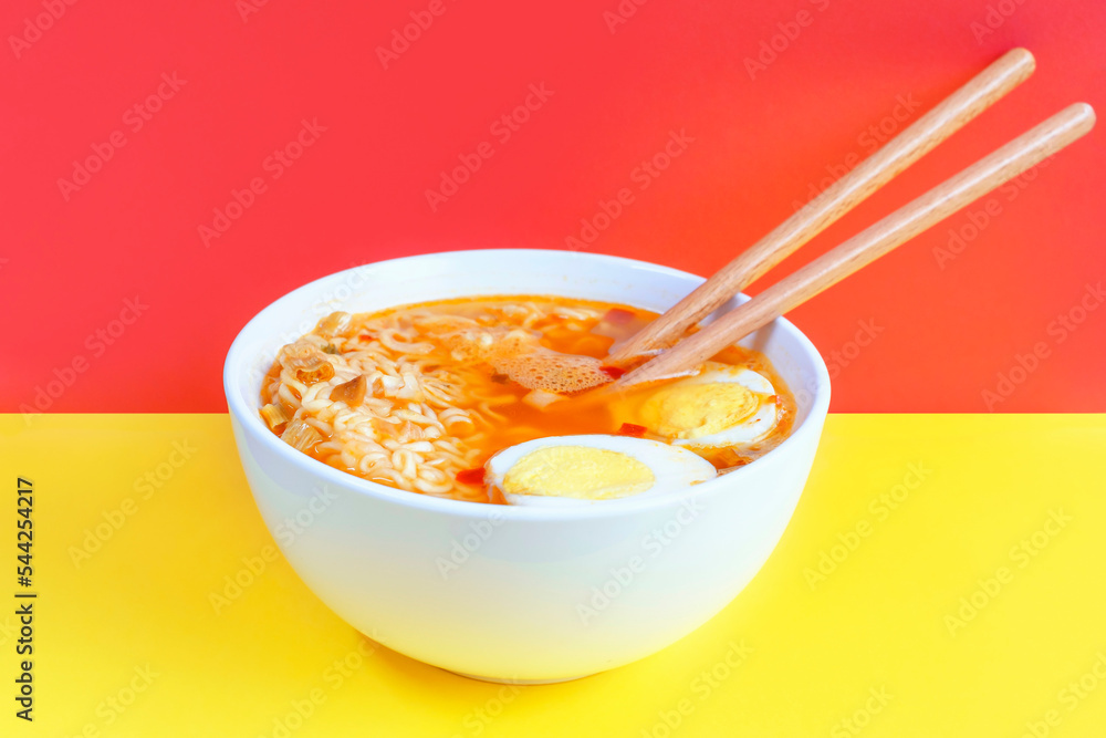 Bowl with instant noodle soup and eggs on red and yellow background close-up, side view. Bowl with instant noodle soup and eggs on red and yellow background close-up, side view.