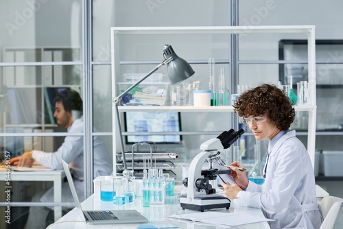 Young chemist or microbiologist in labcoat and protective eyeglasses sitting by workplace with microscope and making notes during trial