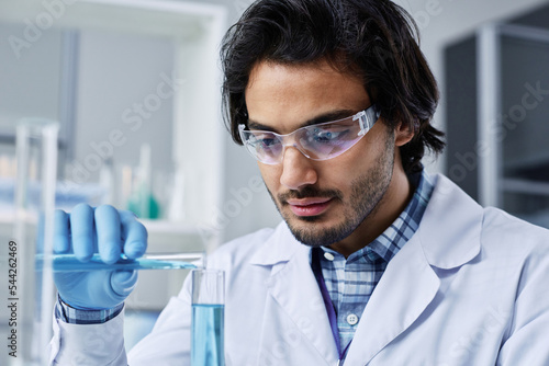 Fototapeta Naklejka Na Ścianę i Meble -  Close-up of young scientist in protective eyeglasses and gloves pouring liquid substance into flask with another one to get new compound