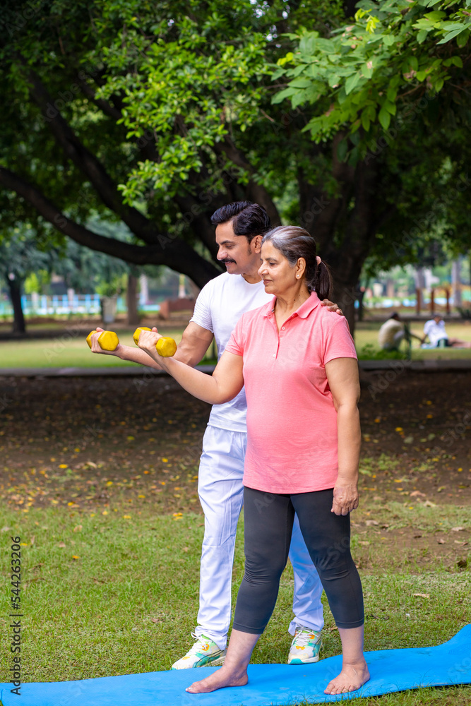 Indian trainer help a woman with dumbbell workout at park, working on arms strength, Asian athletic lady exercising outdoor with coach in nature, fitness concept. Indian trainer help a woman with dumbbell workout at park, working on arms strength, Asian athletic lady exercising outdoor with coach in nature, fitness concept.