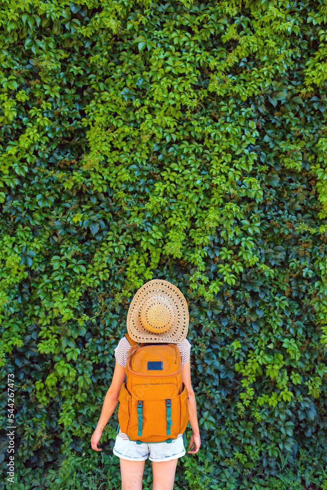 A girl in a straw hat stands near a wall overgrown with ivy