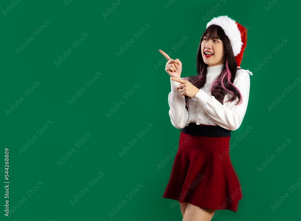 Young pretty asian woman in white and red christmas theme costume wearing santa hat finger pointing for copy space green screen background. Merry Christmas. Young pretty asian woman in white and red christmas theme costume wearing santa hat finger pointing for copy space green screen background. Merry Christmas.