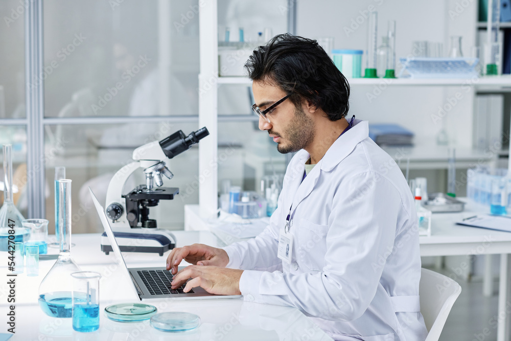 Side view of young serious laboratory worker in labcoat sitting by workplace in front of laptop and searching for online information Side view of young serious laboratory worker in labcoat sitting by workplace in front of laptop and searching for online information
