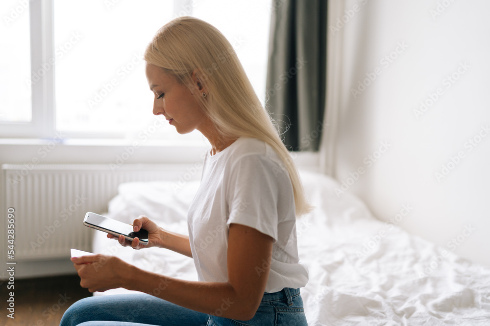 Side view of pretty blonde young woman entering credit card number for online shopping using mobile phone sitting in light room on background of window. Concept of e-commerce user. Side view of pretty blonde young woman entering credit card number for online shopping using mobile phone sitting in light room on background of window. Concept of e-commerce user.