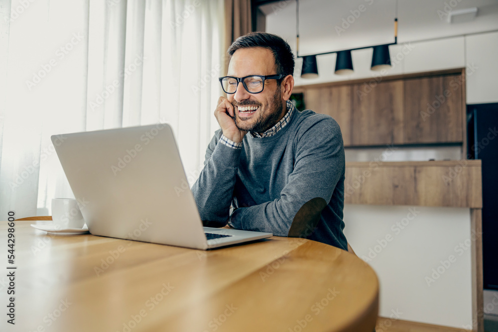 A smiling casual businessman is reading e-mails on laptop while working remotely from home. A smiling casual businessman is reading e-mails on laptop while working remotely from home.
