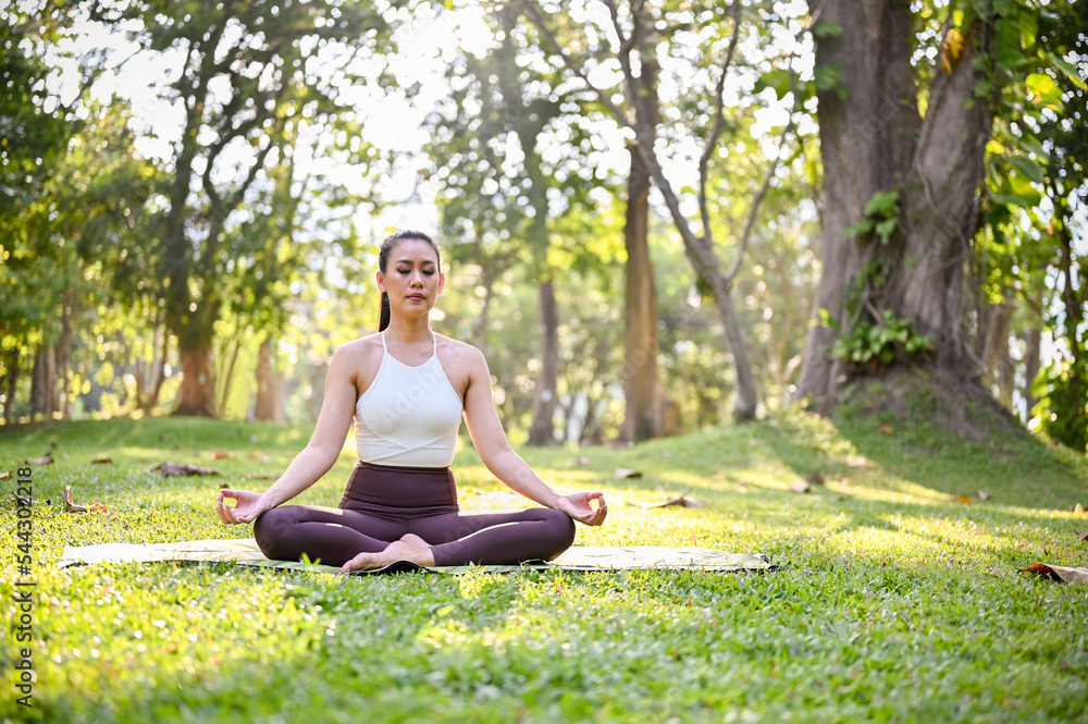 Charming Asian woman sitting on her yoga mat in the garden, focusing on her breath, meditating Charming Asian woman sitting on her yoga mat in the garden, focusing on her breath, meditating