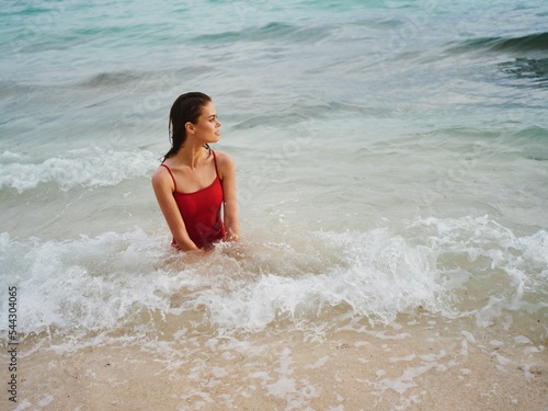 Sexy woman in a red swimsuit with an athletic body stands in the water in the ocean and poses looking out at the horizon