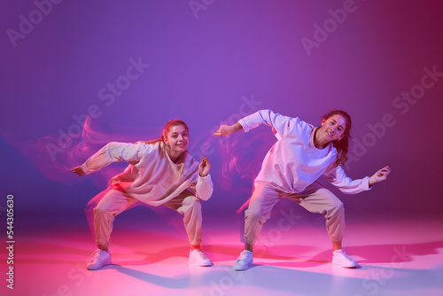 Portrait of two young girls dancing hip-hop isolated over gradient purple background in neon with mixed light © Lustre