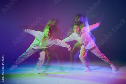Portrait of young girls dancing hip-hop isolated over gradient blue purple background in neon with mixed light. Friendship