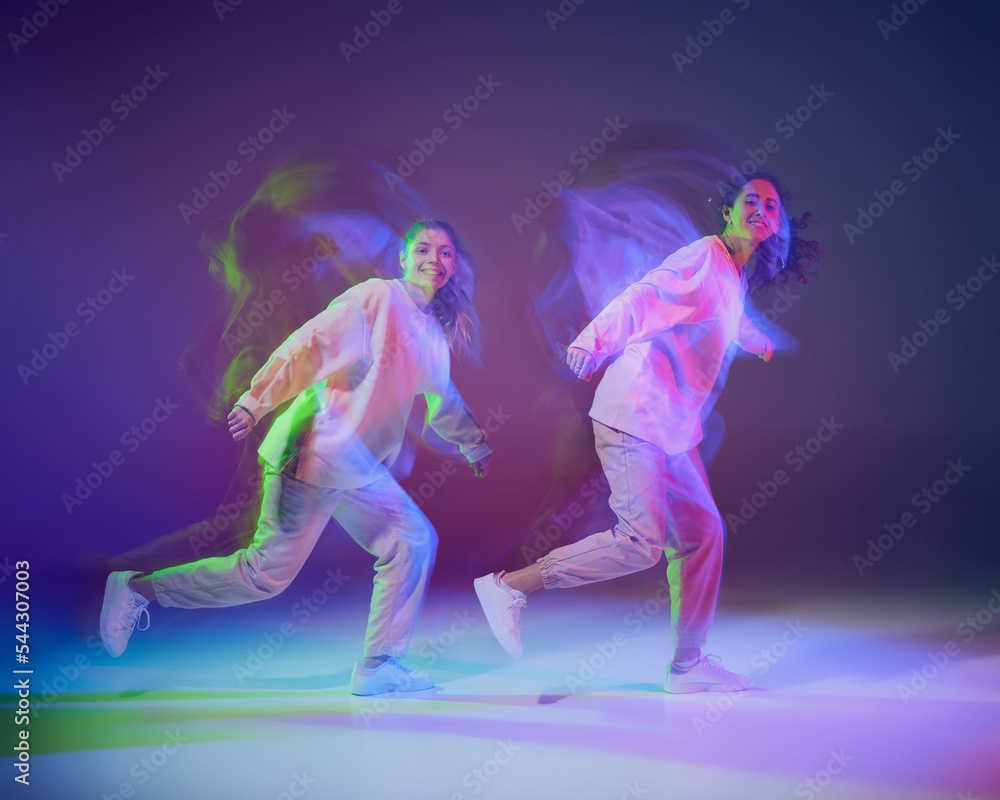 Portrait of young girls dancing hip-hop isolated over gradient blue purple background in neon with mixed light. Looking happy