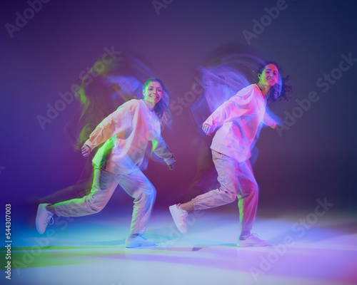 Portrait of young girls dancing hip-hop isolated over gradient blue purple background in neon with mixed light. Looking happy © Lustre