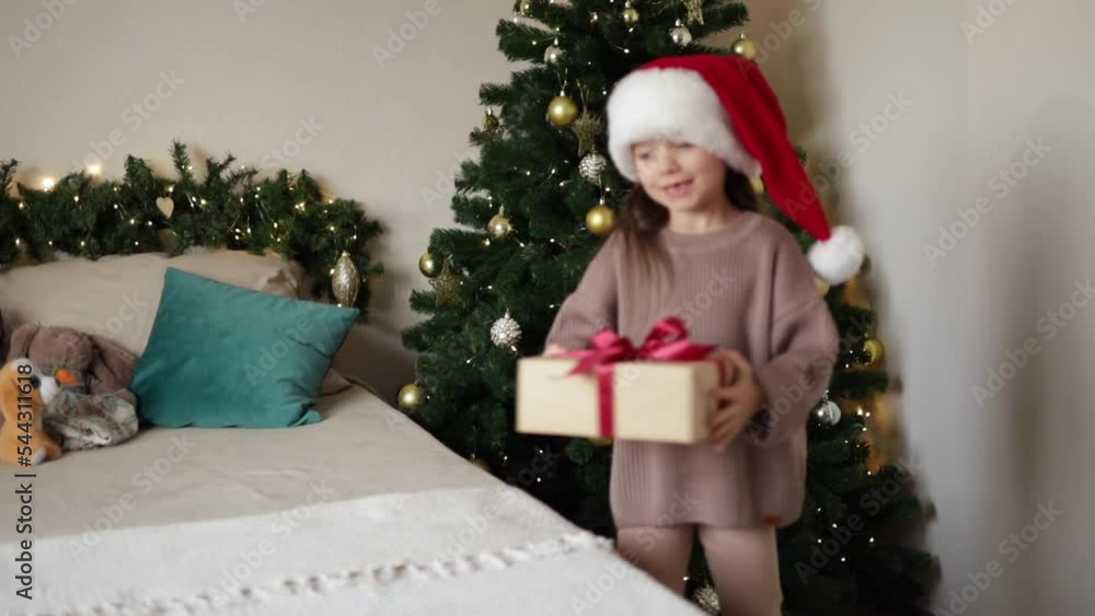 Merry Christmas. A cute cheerful girl in Santa's cap receives a gift near a decorated Christmas tree and opens it. Lifestyle. Natural colours Merry Christmas. A cute cheerful girl in Santa's cap receives a gift near a decorated Christmas tree and opens it. Lifestyle. Natural colours