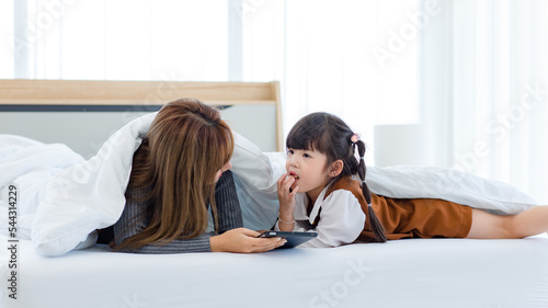 Millennial Asian young pretty female teenager mother nanny babysitter in casual outfit laying down on bed smiling thinking with little cute preschooler thoughtful daughter girl in bedroom in morning
