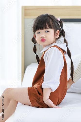 Closeup shot of Asian young little cute preschooler daughter girl in casual dress sitting smiling on cozy bed in bedroom at home. Small tie hairstyle child with brown outfit on weekend in hotel