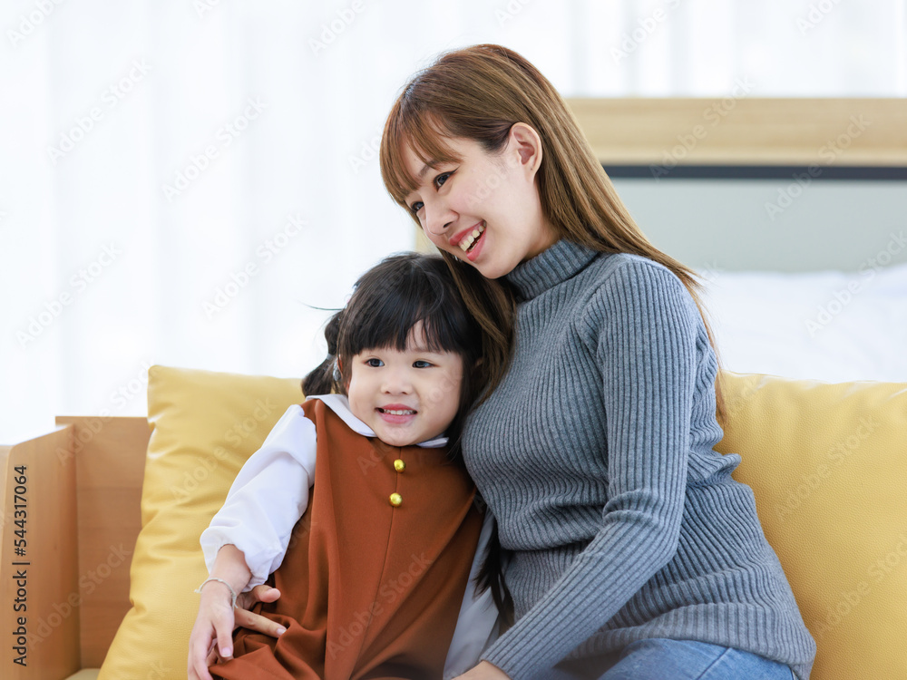 Millennial Asian cheerful happy young pretty female teenager mother nanny babysitter in casual outfit sitting on cozy sofa smiling hugging showing love with little cute preschooler daughter girl Millennial Asian cheerful happy young pretty female teenager mother nanny babysitter in casual outfit sitting on cozy sofa smiling hugging showing love with little cute preschooler daughter girl