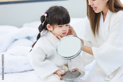 Closeup shot of Millennial Asian happy cheerful little cute preschooler daughter girl wearing white bathrobe after bath looking to mirror in hands setting hairstyle with unrecognizable unknown mother