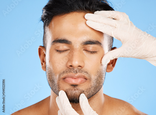 Cosmetic, plastic surgery and face of young man, for skincare and white gloves on blue studio background for dermatology care. Makeup, Indian male and skin rejuvenation for wellness and collagen.