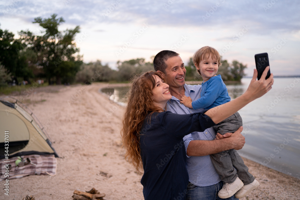 Fototapeta premium Portrait of Caucasian family with father, mother and little son selfie with lake view by smartphone while vacation together in holiday. Happy family time