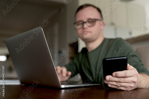 Low angle of adult caucasian man with down syndrome using laptop and phone at home