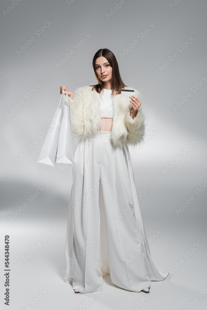 Full length of trendy woman in white clothes holding credit card and shopping bags on abstract grey background. Full length of trendy woman in white clothes holding credit card and shopping bags on abstract grey background.
