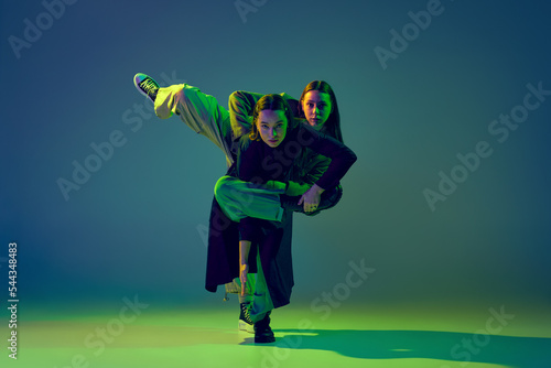 Stylish young girls dancing casual clothes dancing experimental style dance over gradient blue-green background at dance hall in neon light. Youth culture photo