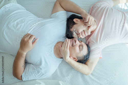 Asian young male gay family lying down on bed and look at each other. Attractive handsome LGBTQ couple in pajamas, feel happy after wake up in morning and kiss forehead of boyfriend in bedroom at home