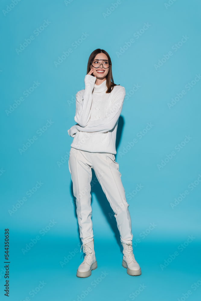 full length of joyful young woman in eyeglasses and totally white outfit posing on blue. full length of joyful young woman in eyeglasses and totally white outfit posing on blue.