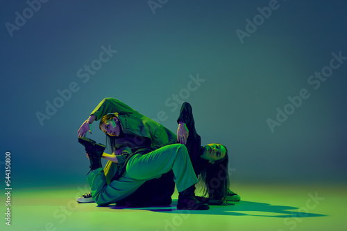 Young stylish flexible girls in modern attires dancing contemp style dance isolated over gradient blue-green background at dance hall in neon light. photo