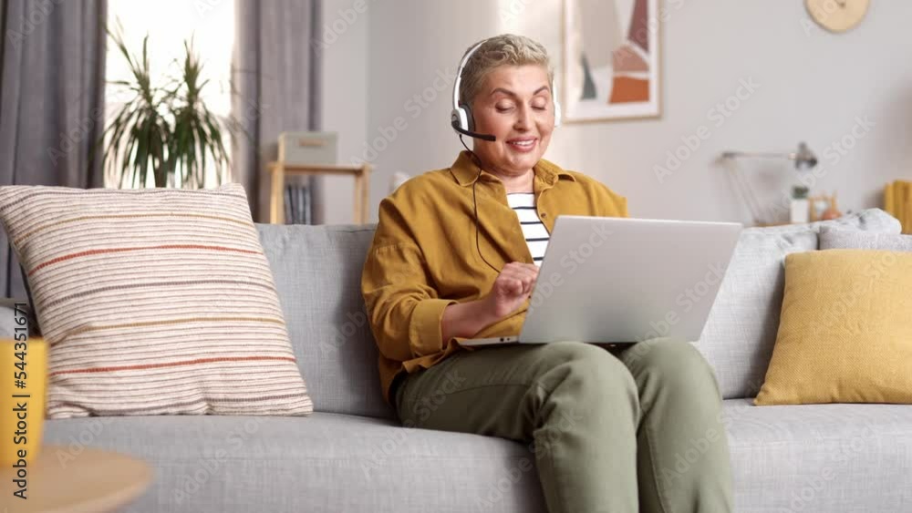 Beautiful cute middle aged woman working from home online on laptop sitting on sofa wearing headphones. Busy concentrated happy short haired female calling customers. Working from home concept. Beautiful cute middle aged woman working from home online on laptop sitting on sofa wearing headphones. Busy concentrated happy short haired female calling customers. Working from home concept.