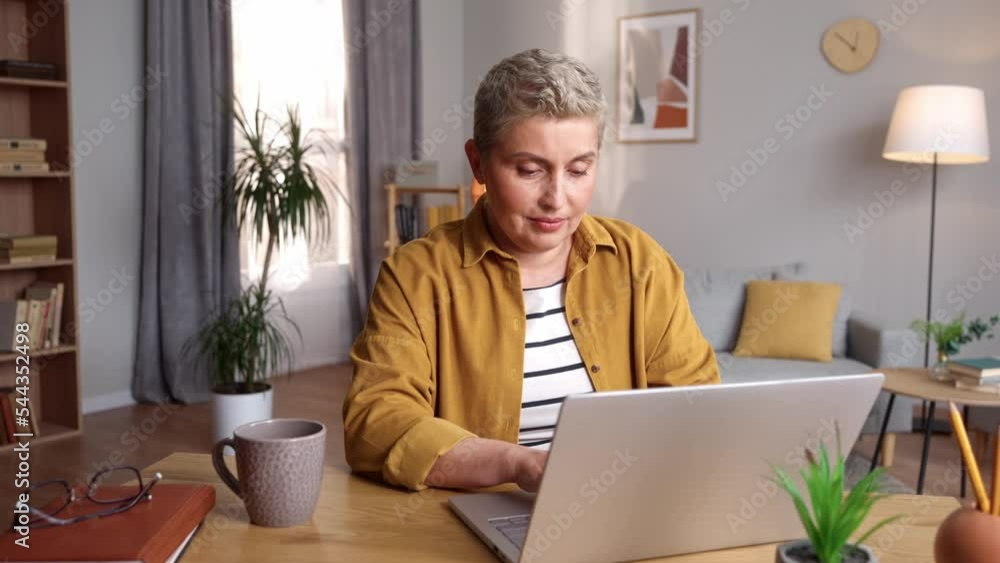 Modern attractive concentrated woman typing on laptop sitting at table in living room. Middle-aged blonde female working from home writing online freelance Internet technologies. Freelance concept Modern attractive concentrated woman typing on laptop sitting at table in living room. Middle-aged blonde female working from home writing online freelance Internet technologies. Freelance concept