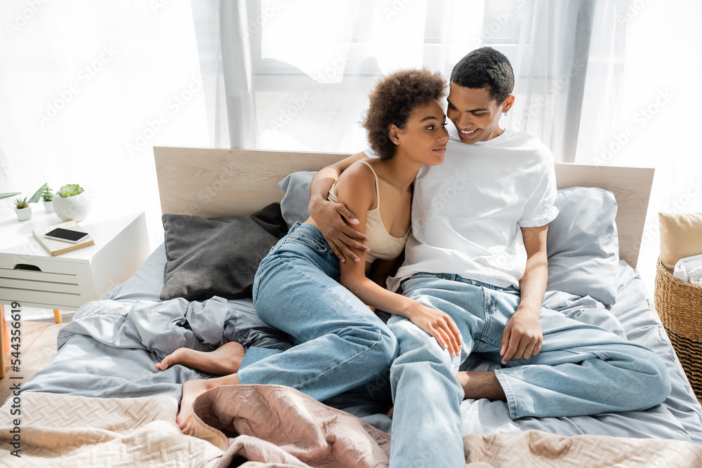 cheerful african american man in jeans and white t-shirt hugging curly girlfriend while sitting on bed.