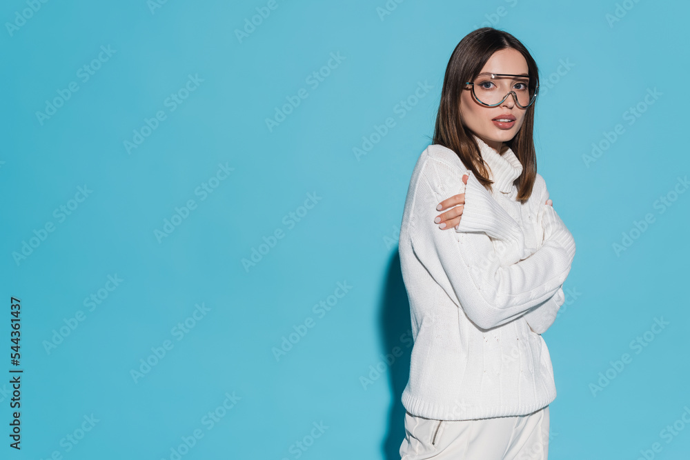 pretty young woman in trendy eyeglasses and totally white outfit posing with crossed arms on blue. pretty young woman in trendy eyeglasses and totally white outfit posing with crossed arms on blue.