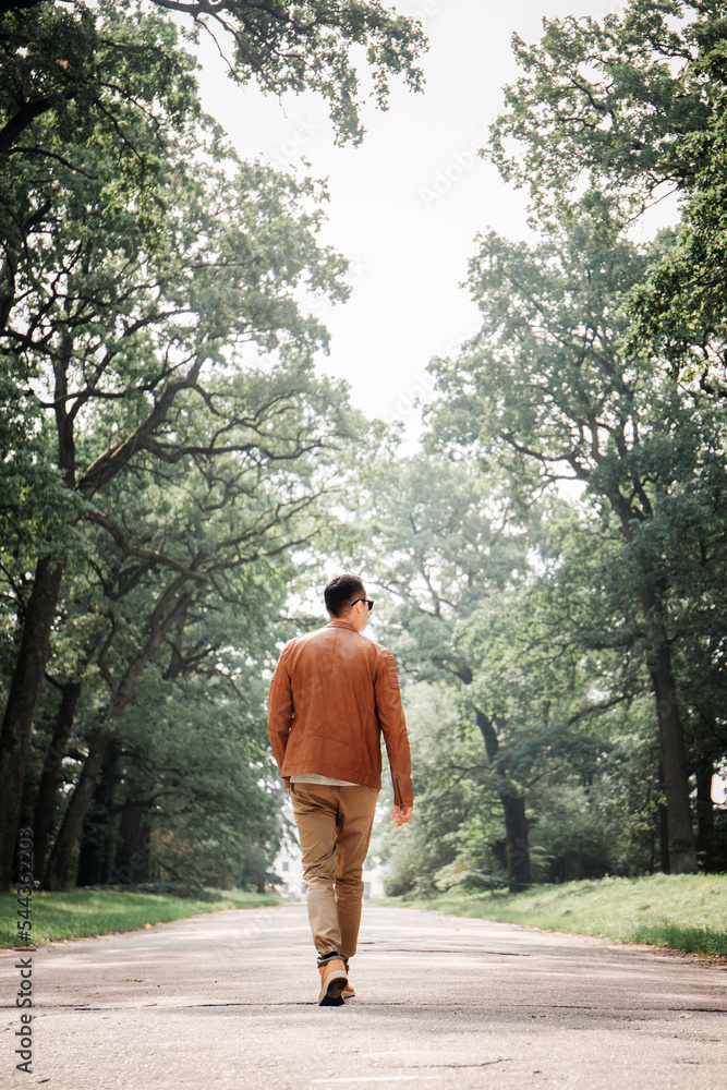 A young handsome guy is walking along a wide road with huge trees, back view