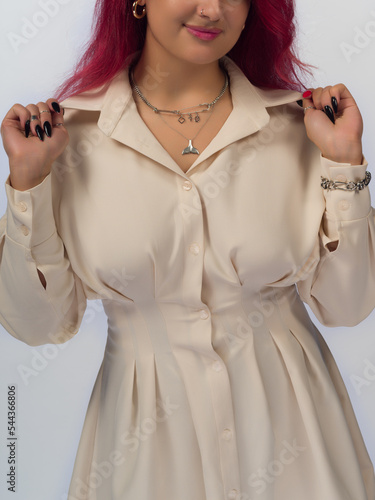A young girl with long brown hair on a white background. She's wearing a beige dress. Wears piercings and jewelry. Close-up of the dress.