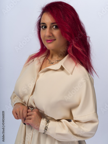 A young girl with long brown hair and brown eyes on a white background. She's wearing a beige dress. Wears piercings and jewelry. Close-up