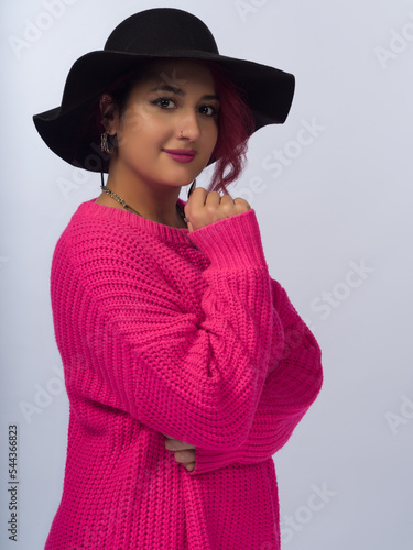A girl with brown hair on a white background. She is wearing a black wide-brimmed hat with a wide brim and a pink loose-fitting jumper. Close-up
