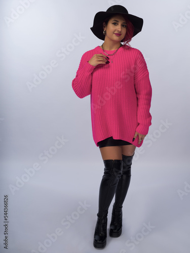 A girl with brown hair on a white background. She is wearing a black wide-brimmed hat with a wide brim and a pink loose-fitting jumper. The skirt is above the knee. Black high boots. Full height.