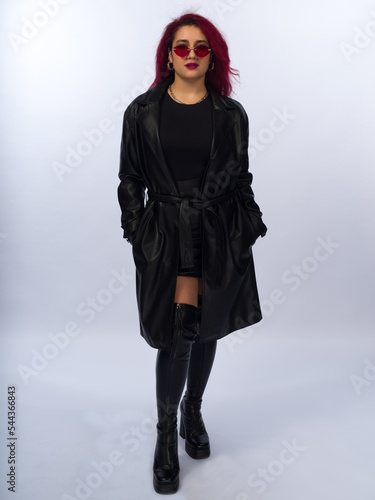 Stylish young girl with brown hair on a white background. She is dressed in a black leather raincoat and a skirt above the knee. Wears sunglasses, piercings. Full-length photo
